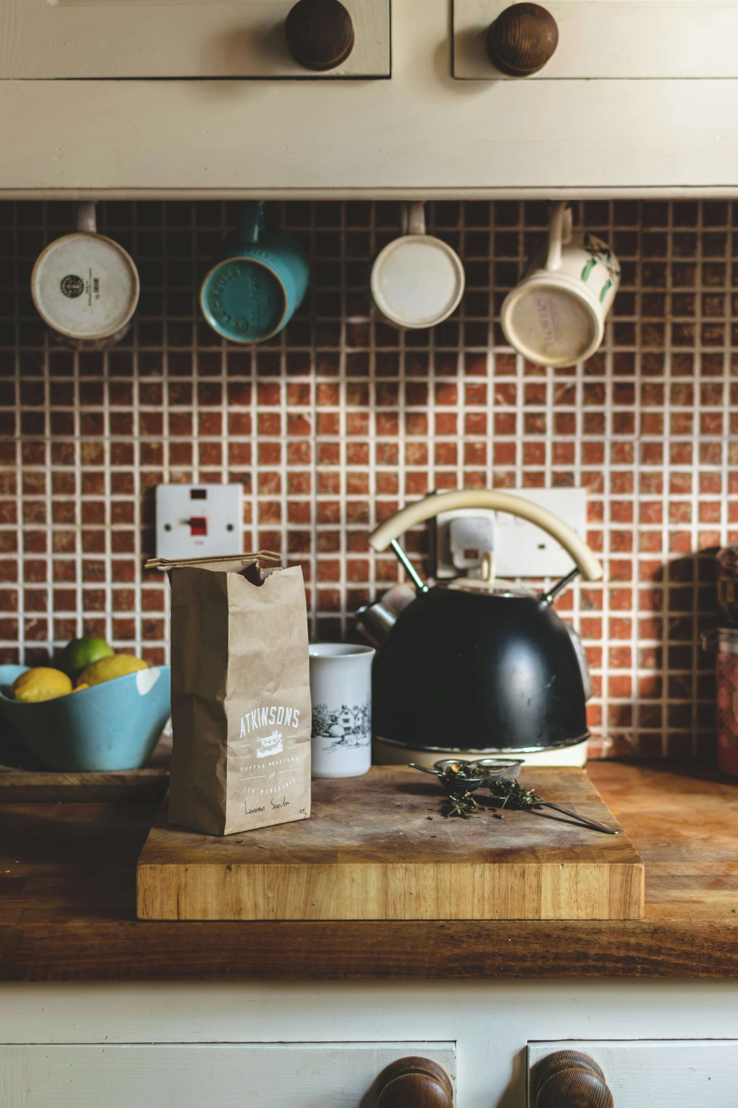 Paquet de café posé sur une planche en bois dans une cuisine vintage, avec une bouilloire noire, des tasses suspendues et un saladier de citrons en arrière-plan.