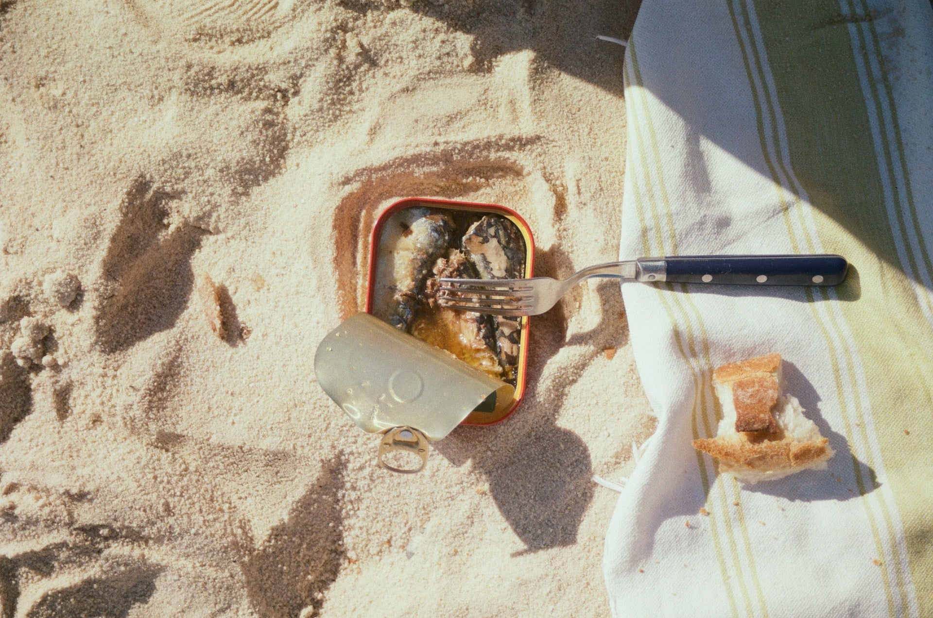 Boîte de sardines ouverte sur le sable, avec une fourchette, un morceau de pain et une serviette de pique-nique.