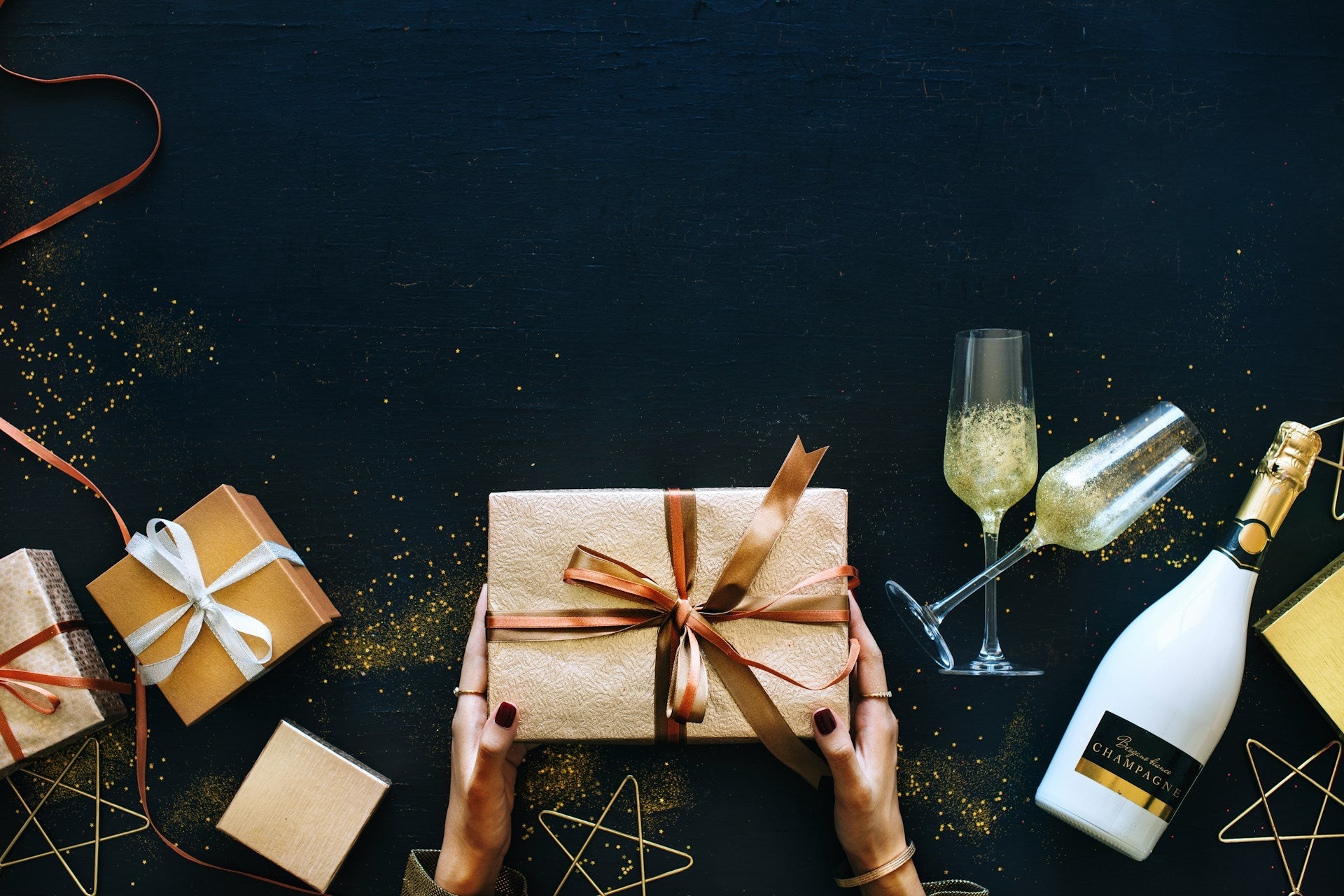 Mains tenant un coffret cadeau doré avec ruban, entouré de paquets, flûtes et bouteille de champagne sur fond noir élégant